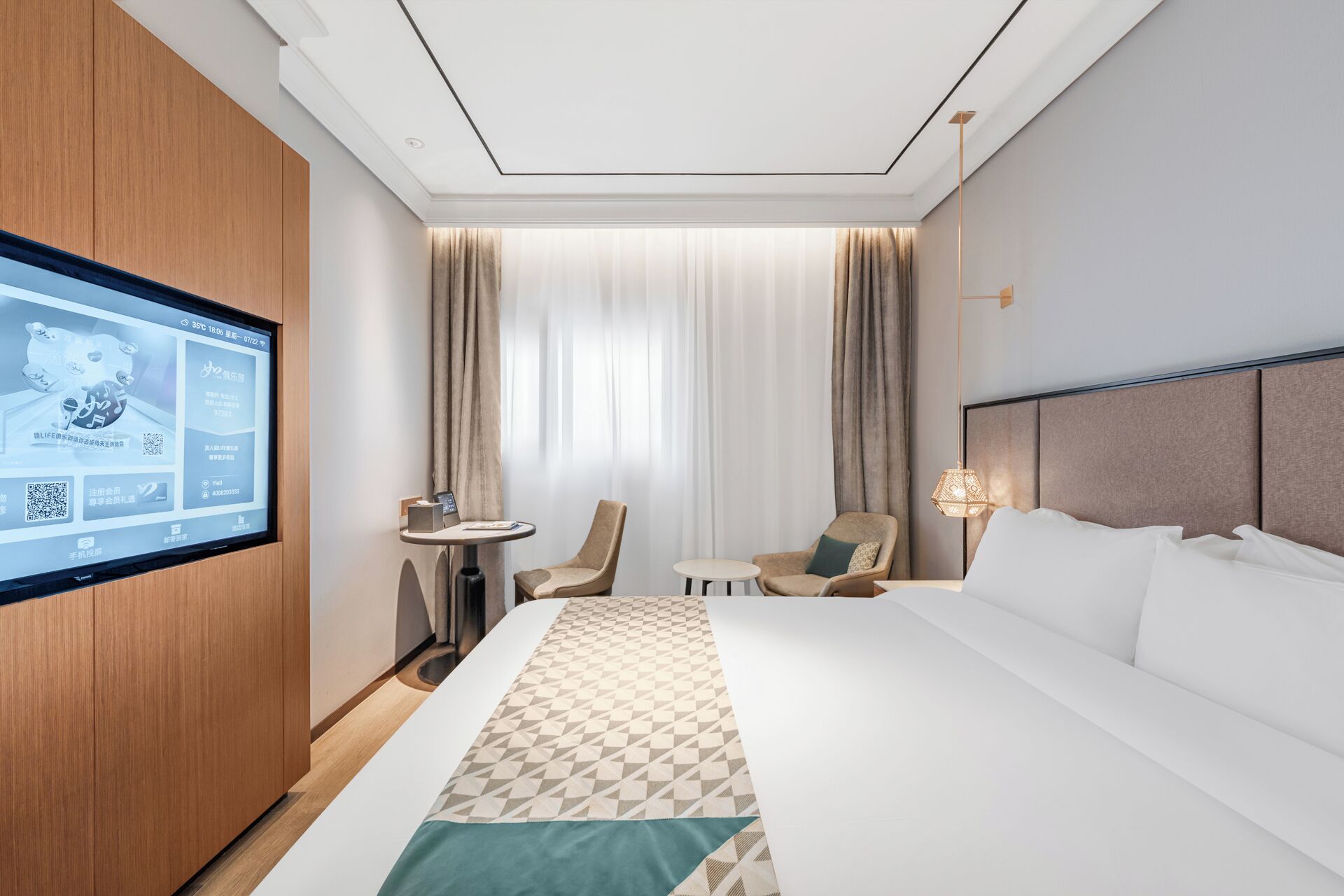 A hotel room with a bed, a TV mounted on the wall, a small table, two chairs, and a window with curtains.