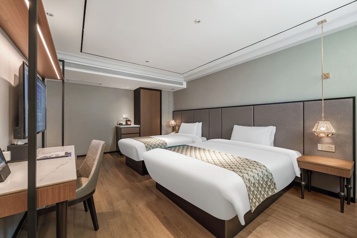A hotel room with two beds, a desk, a chair, a TV, a minibar, and a bedside table with a lamp.