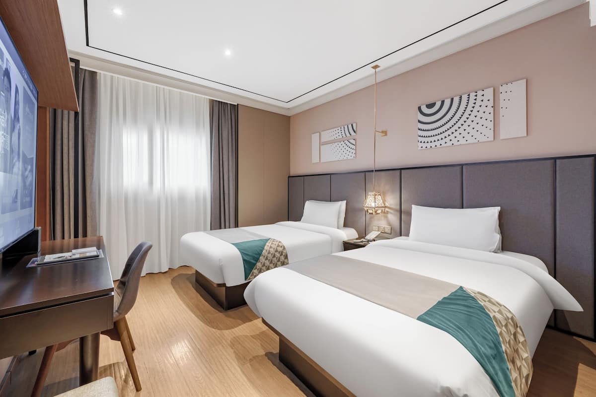 A hotel room with two beds, a desk, a TV, and a window with curtains.