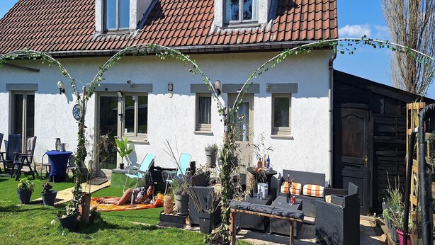 Een huis met twee verdiepingen, voorzien van een pannendak, een klein raam en een deur. Er zijn zitgedeeltes buiten met stoelen en tafels, en er staan potten met planten rondom.