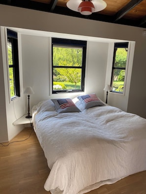 A bedroom with a bed, two windows, a lamp, and a view of greenery.
