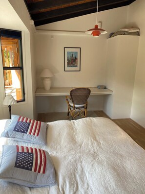 Ein Schlafzimmer mit einem Bett, zwei Kissen, einem Sessel, einem Schreibtisch, einer Lampe, einem Bild an der Wand und einem Fenster.
