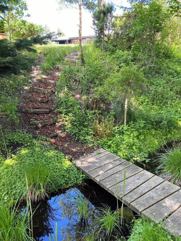 Eine Holzbrücke über ein kleines Gewässer, umgeben von üppiger Vegetation und einem Pfad zu einem Gebäude.