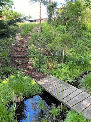 A wooden bridge over a small water body, surrounded by lush greenery and a path leading to a building.