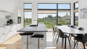 Une cuisine moderne dotée d’un grand îlot central, d’un coin repas avec des chaises et de grandes fenêtres offrant une vue sur la verdure.