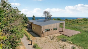 Une cabane moderne en bois, dotée d’un toit plat, de grandes fenêtres et d’une terrasse, qui offre une vue sur une zone verdoyante et la mer.