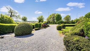 Un chemin de gravier bordé de haies et d’arbres soigneusement taillés, qui mène à un point d’observation offrant une vue lointaine sur le paysage.