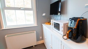 Een kleine kitchenette met een wit aanrecht, een magnetron, een koffiezetapparaat, een aan de muur bevestigde televisie en een raam met jaloezieën.