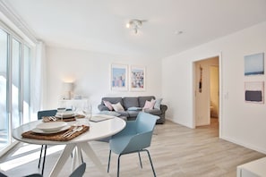 Un salon moderne avec une table à manger pour deux personnes, un canapé gris et un fauteuil bleu. Deux œuvres d’art encadrées ornent le mur, ainsi qu’un luminaire au plafond et une porte qui donne sur une autre pièce.