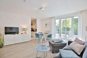 A modern living room with a flat-screen TV, a dining area with blue chairs, and a balcony with outdoor furniture. - Bergstraße 62 Vacation Home, Apartment 5, Tide am Park - Bergstraße 62 Residence (Timmendorfer Strand)