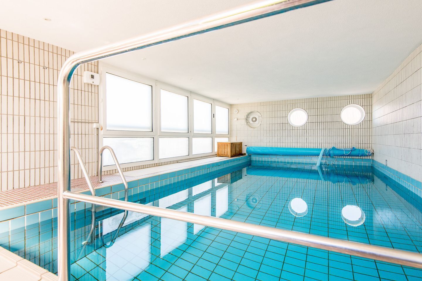 Une piscine aux carreaux bleu clair, une échelle en métal et un mur vitré.