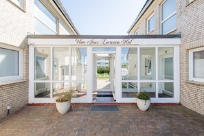 A building entrance with a sign reading 'Ulve Jens Lornsen-Hof,' large glass windows, and two potted plants on the ground. - Lornsen-Hof, Apt. 2 - Modern ground-floor apartment with 2 rooms, approximately 50 m², (Sylt)
