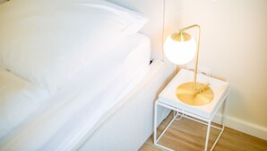 Un lit avec une literie blanche, une table de chevet avec une lampe et un plancher en bois.