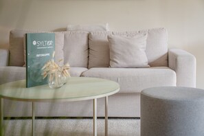 A living room with a sofa, a glass coffee table, and a book titled 'SYLTEN INFOMAPPE'. - Lornsen-Hof, App. 2 - Modern apartment on the ground floor with 2 rooms on approx. 50 m², (Sylt)