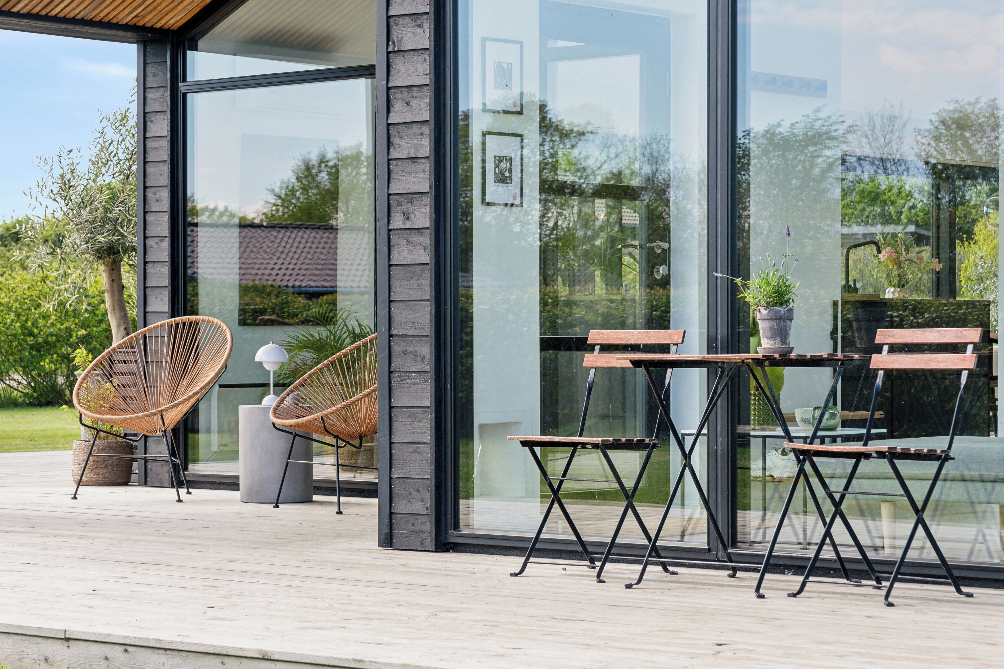 Una terraza moderna al aire libre con sillas de mimbre, una mesa de madera, ventanas de vidrio amplias y vistas a zonas verdes.
