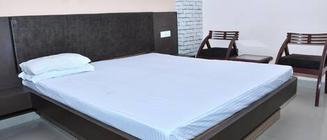 Una cama bien hecha con colchón y almohadas blancas, cabecera oscura y una pequeña mesa con sillas.