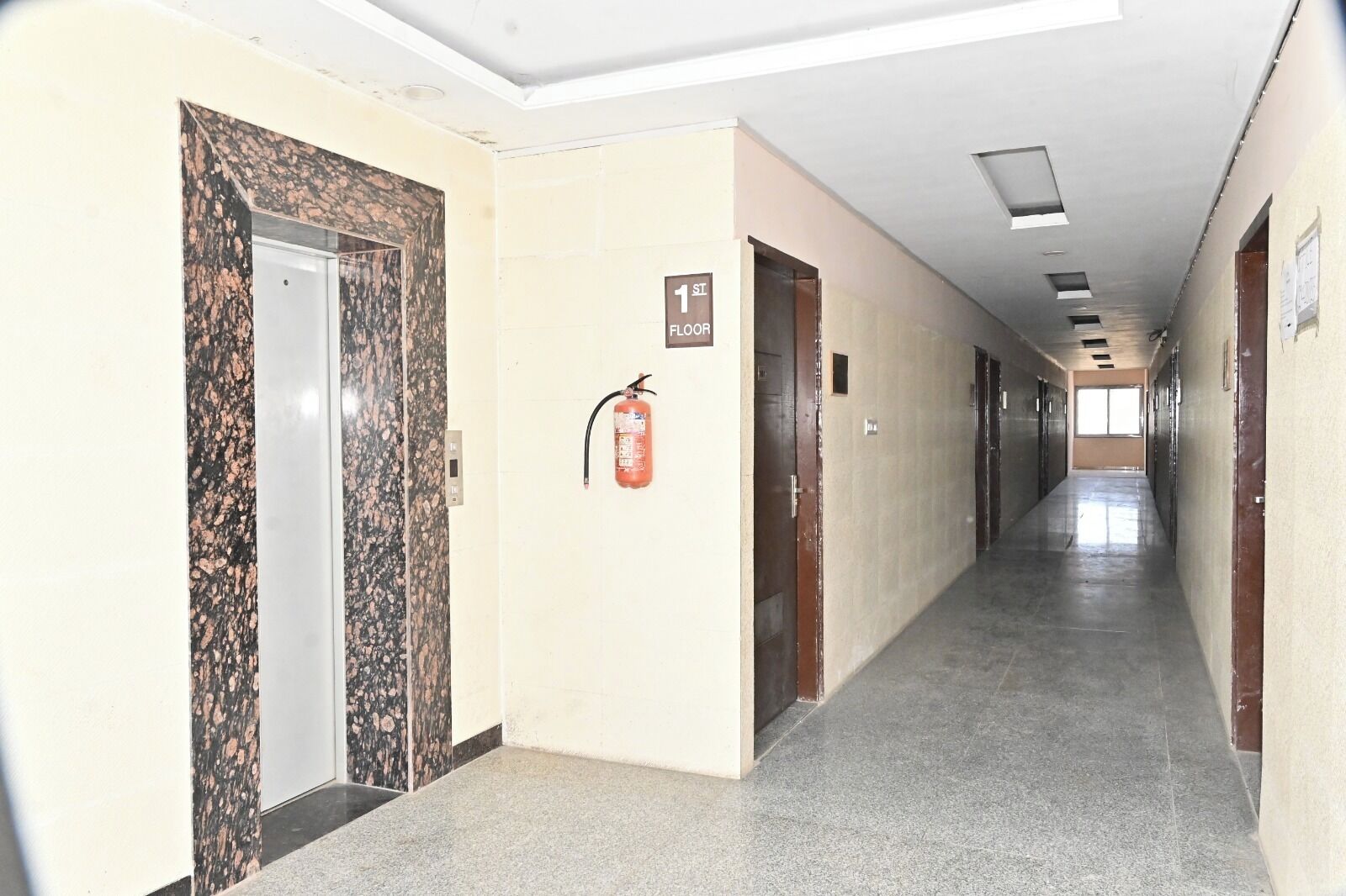 A hallway with a fire extinguisher mounted on the wall, a door with a number sign, and a ceiling with recessed lighting.