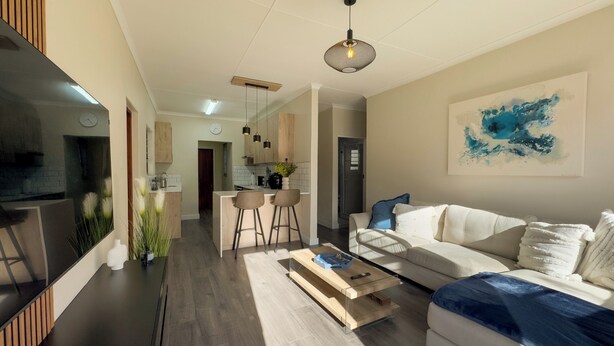 Un salon moderne avec un canapé, une table basse et un téléviseur. On aperçoit à l’arrière-plan un coin cuisine avec une table et des chaises.
