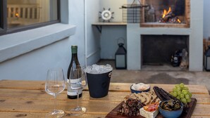 Une table en bois avec une bouteille de vin, un verre et un seau à glace. On y trouve également divers amuse-gueules et un bol d’olives.