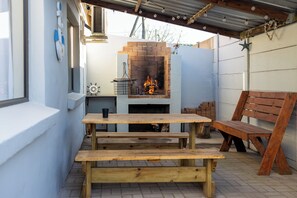 Um pátio externo com lareira de tijolos, bancos de madeira e uma mesa rústica de madeira.