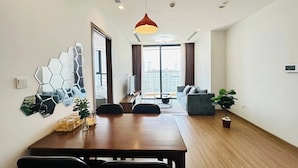 A modern living room with a wooden dining table, a sofa, and a large window offering a city view. - Vinhome Sky Lake-Sunhometel (Hanoi)