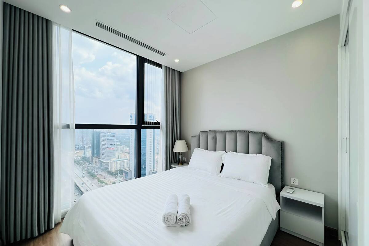 Une chambre d’hôtel moderne avec un grand lit, une table de chevet, une lampe et une vue sur la ville grâce à de grandes fenêtres.