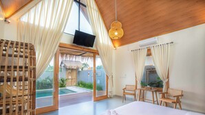A hotel room with a large bed, a television, a dining area with chairs, and a view of a pool and garden. - Villa Norte (Ngaglik)