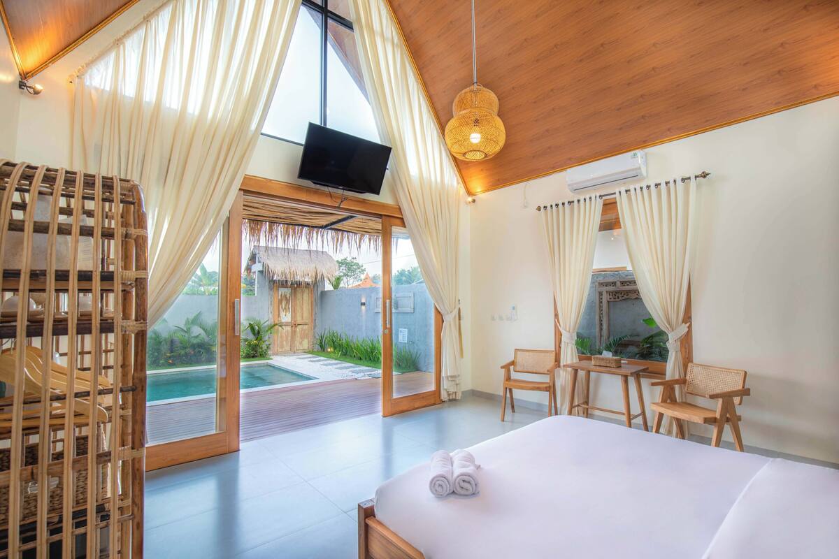 A hotel room with a large bed, a television, a dining area with chairs, and a view of a pool and garden.