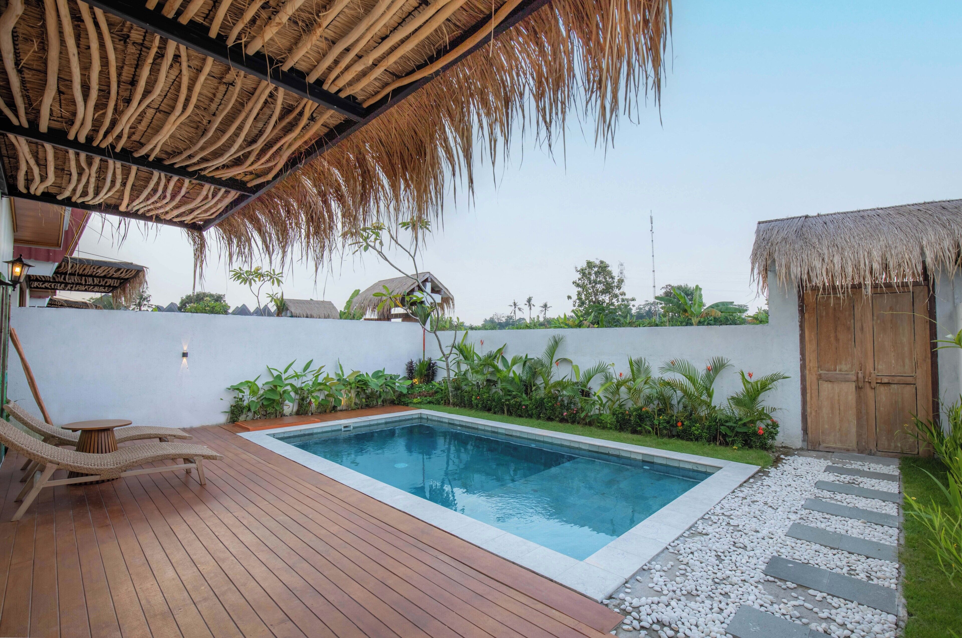 Un área de piscina con maderas, sillones de descanso, techo de paja y pared blanca con puerta de madera.