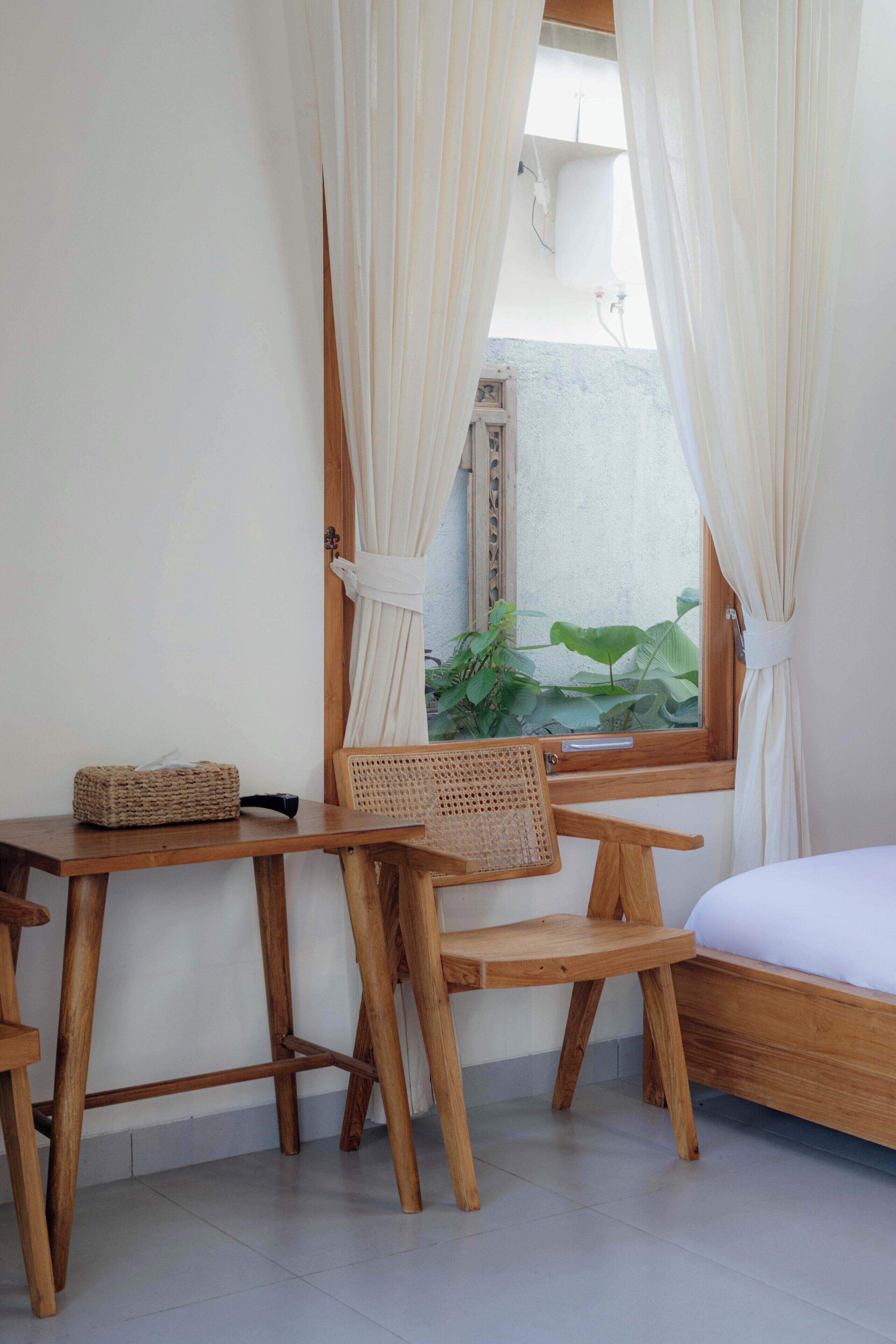 A room with a wooden bed, a small table with a woven basket, and a chair. There is a window with curtains and a view of greenery outside.