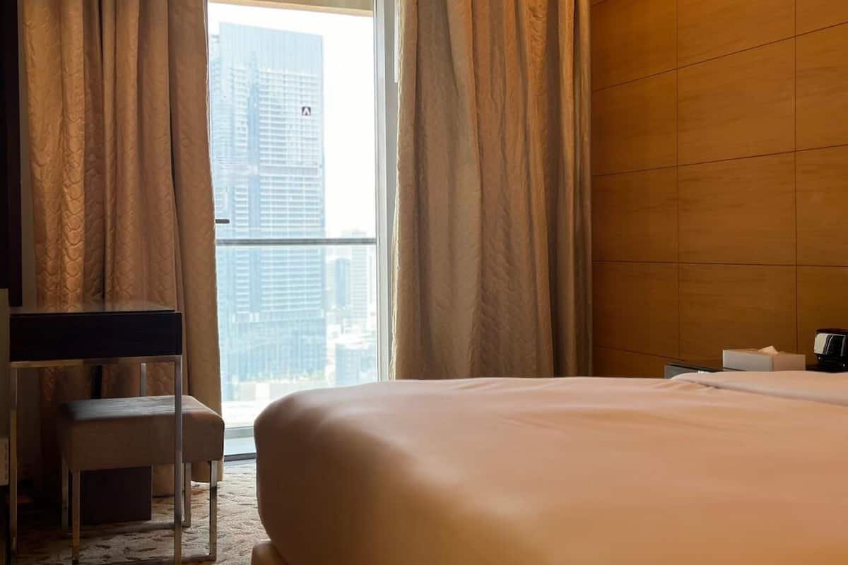 A hotel room with a large bed, a desk, and a view of a cityscape through sheer curtains.