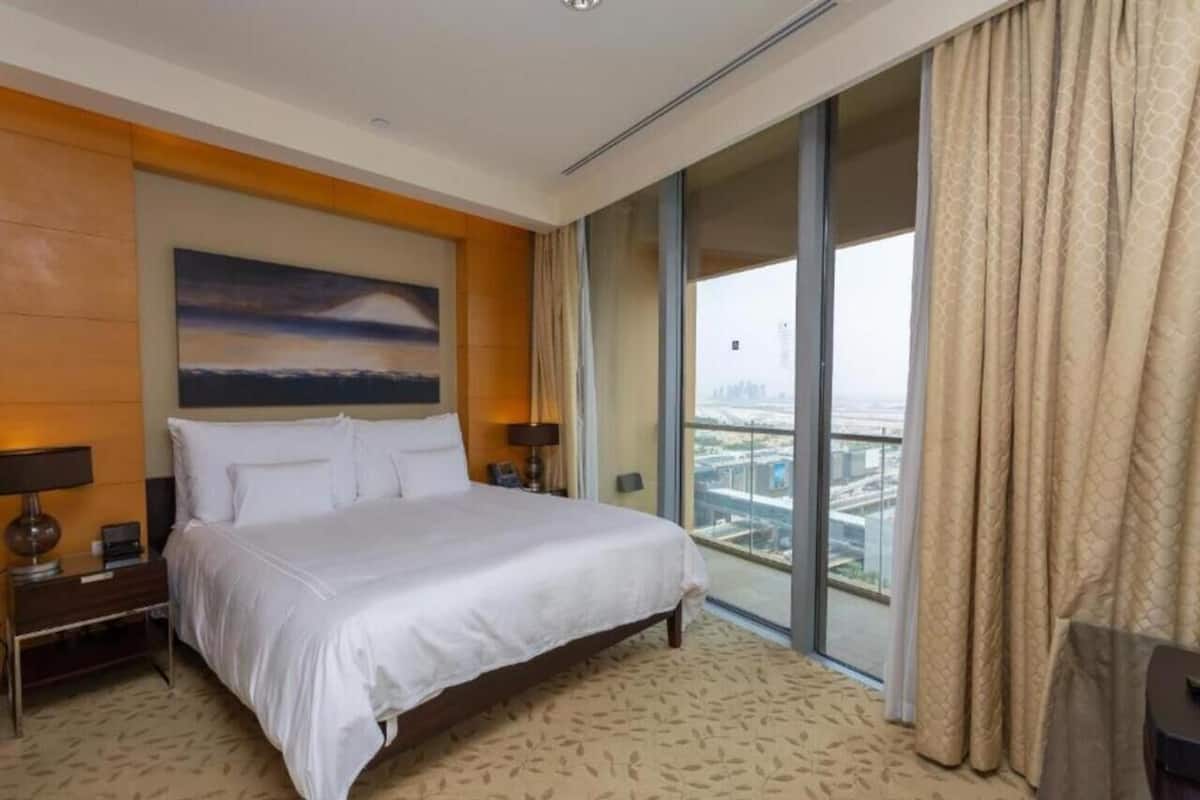 A hotel room with a large bed, a nightstand, a lamp, a painting on the wall, and a view of a cityscape through a large window.