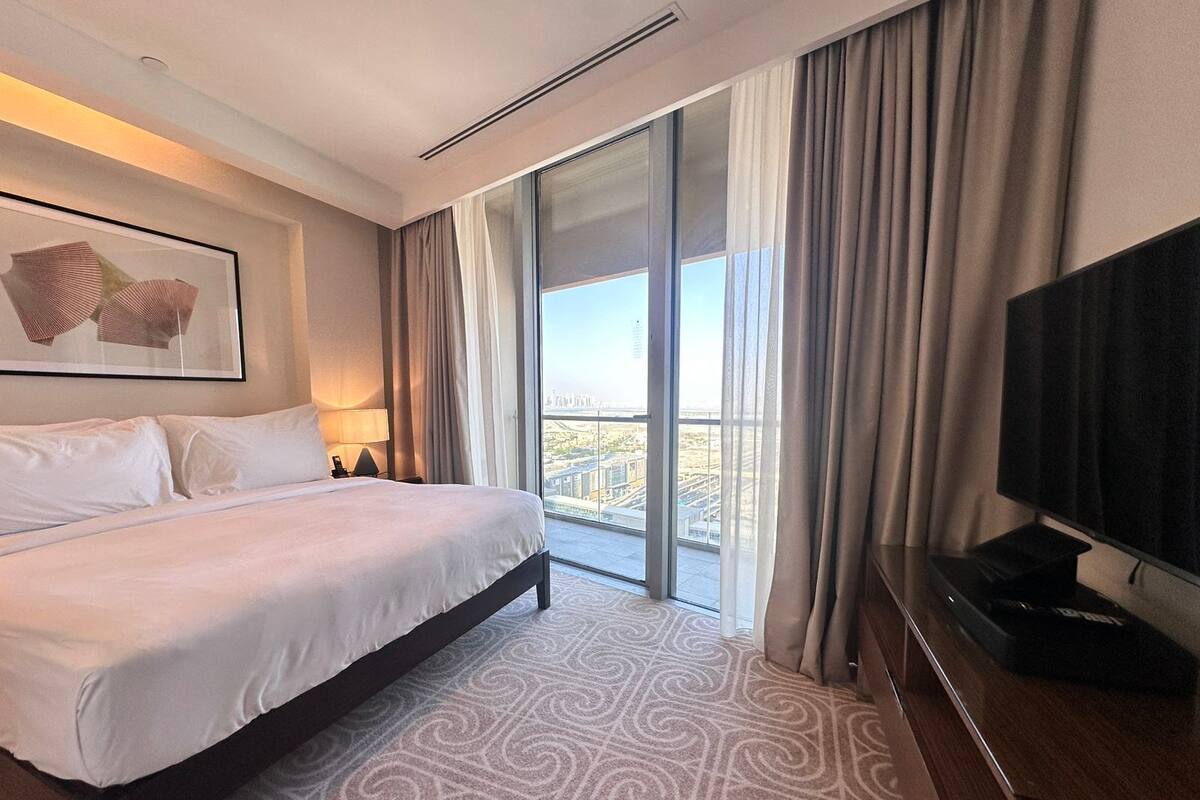 A hotel room with a large bed, a flat-screen TV, a large window with curtains, and a city view.