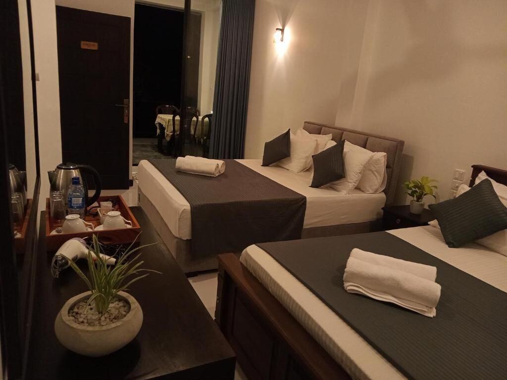 A hotel room with two beds, a small table with a kettle and a tray, a potted plant, and a nightstand with a lamp.
