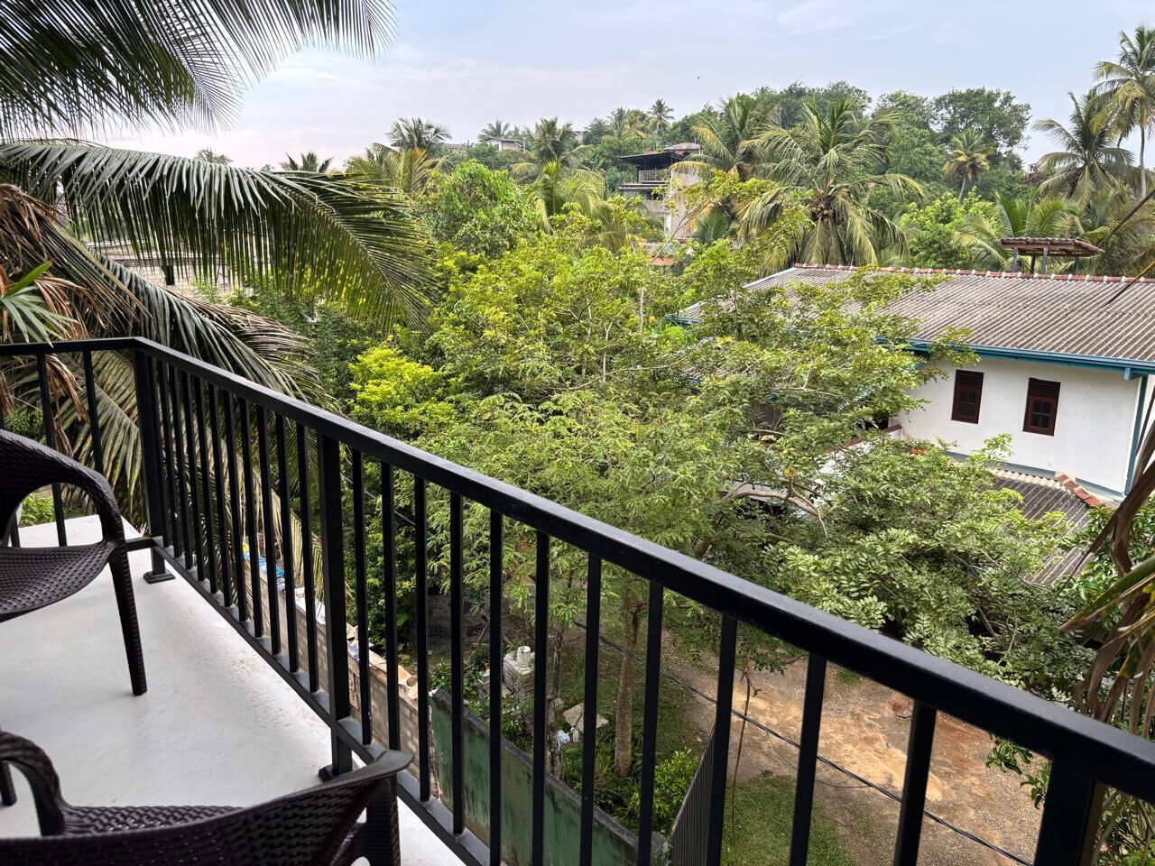 A balcony with a view of a lush garden and a white building with a red roof.