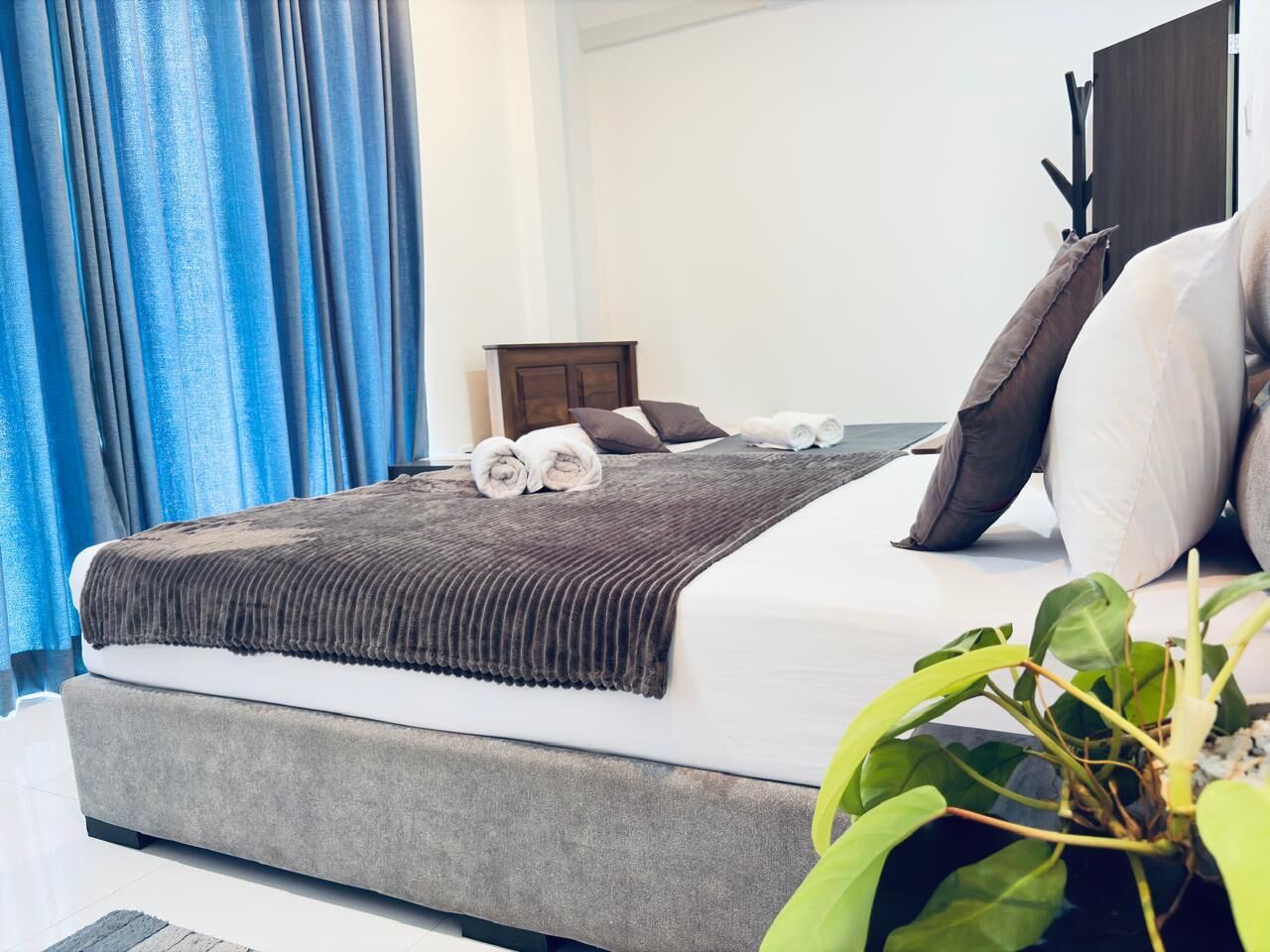 A neatly made bed with a grey upholstered headboard, a brown throw blanket, and rolled towels.