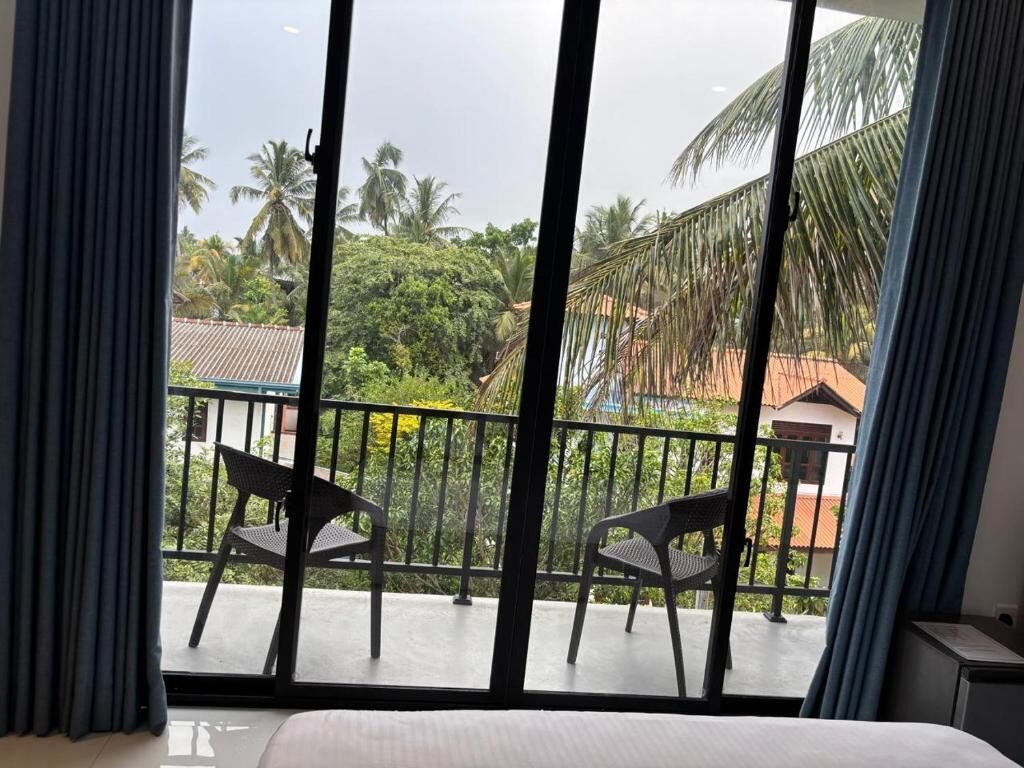 A room with a large window, a bed, and two chairs on a balcony with a view of palm trees and buildings.