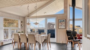 Un vaste espace dédié à la salle à manger, avec une grande table et des chaises, de grandes fenêtres offrant une vue sur l’extérieur, et un coin salon confortable agrémenté d’une cheminée.