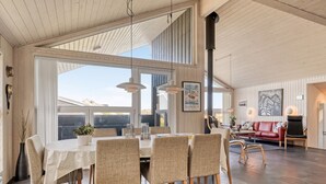 Une salle à manger spacieuse avec un haut plafond et de grandes fenêtres, un salon confortable avec un canapé rouge et, au fond, une cuisine moderne.