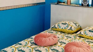 Um quarto com parede azul, uma cama com colcha florida, duas almofadas cor de rosa, uma pequena prateleira com abajur e um letreiro 'Garage' na parede.