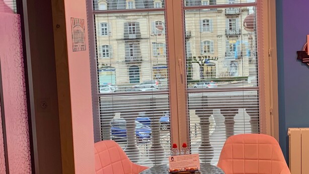 Quarto com duas cadeiras cor-de-rosa e uma pequena mesa, janela com vista para edifícios antigos e parede roxa.
