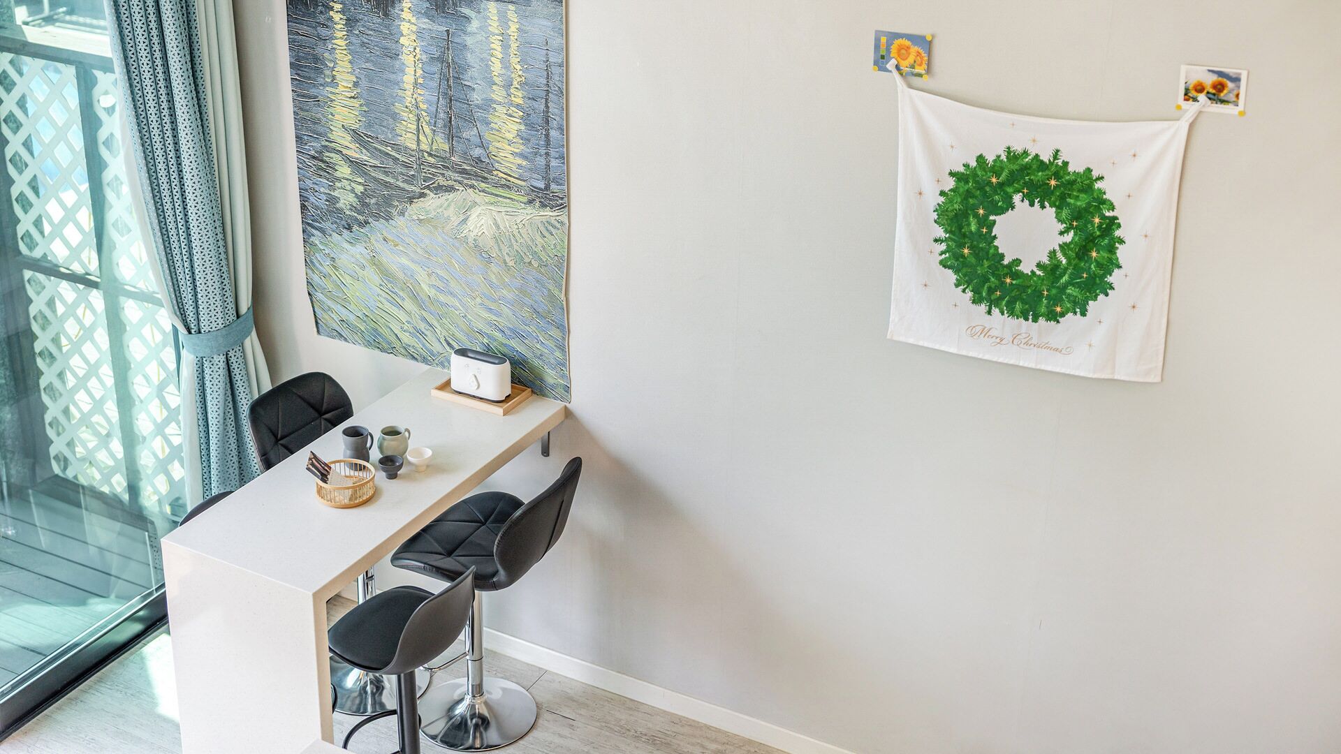 A dining area with a white table, two black chairs, and a green wreath with 'Merry Christmas' on a wall.