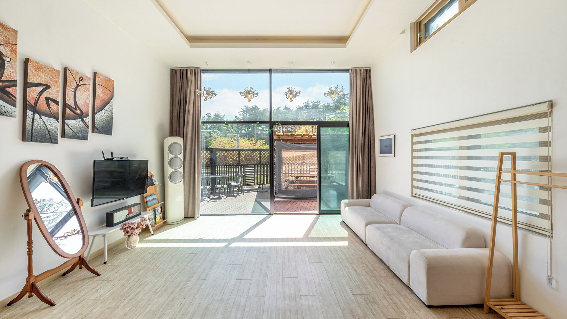 A modern living room with a flat-screen TV, a sofa, a wooden floor, and a balcony with a view of greenery.