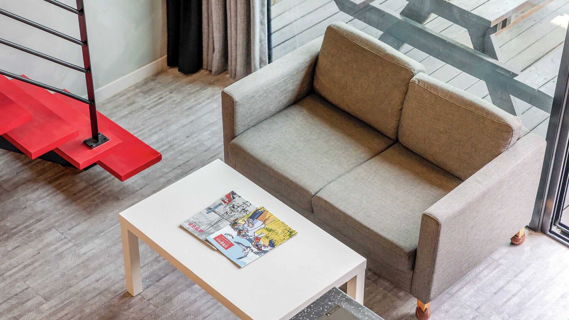 A modern living room with a grey sofa, a white coffee table with magazines, and a red staircase.