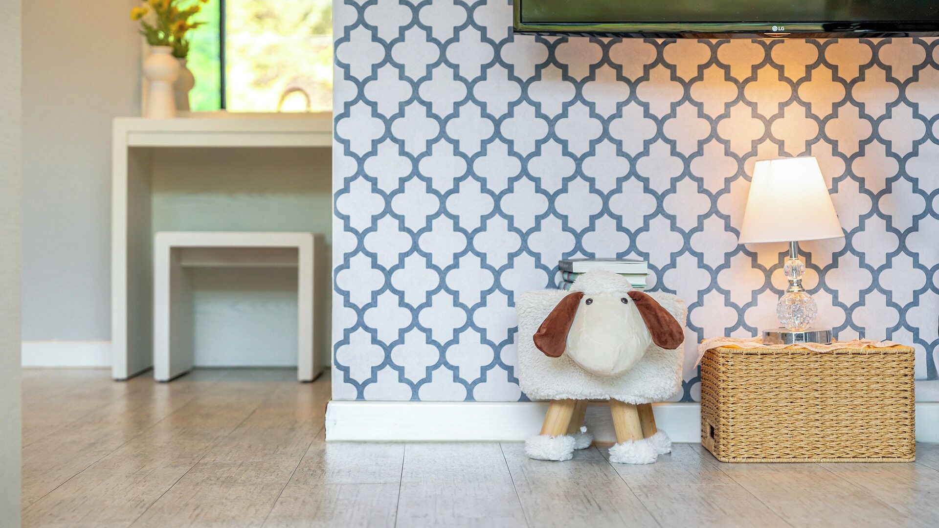 A room with a patterned wall, a television mounted above a white console table, a plush toy on a small stool, and a lamp on a wicker side table.