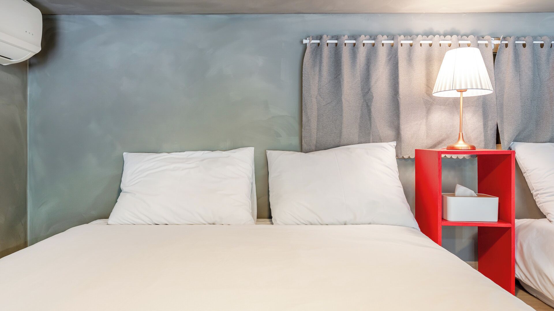 A hotel room with a bed, two pillows, a nightstand with a lamp, and a curtain.