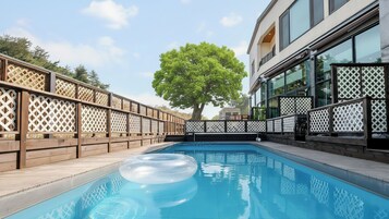 Una piscina con cubierta de madera, un árbol grande al fondo y un edificio moderno con ventanas amplias.