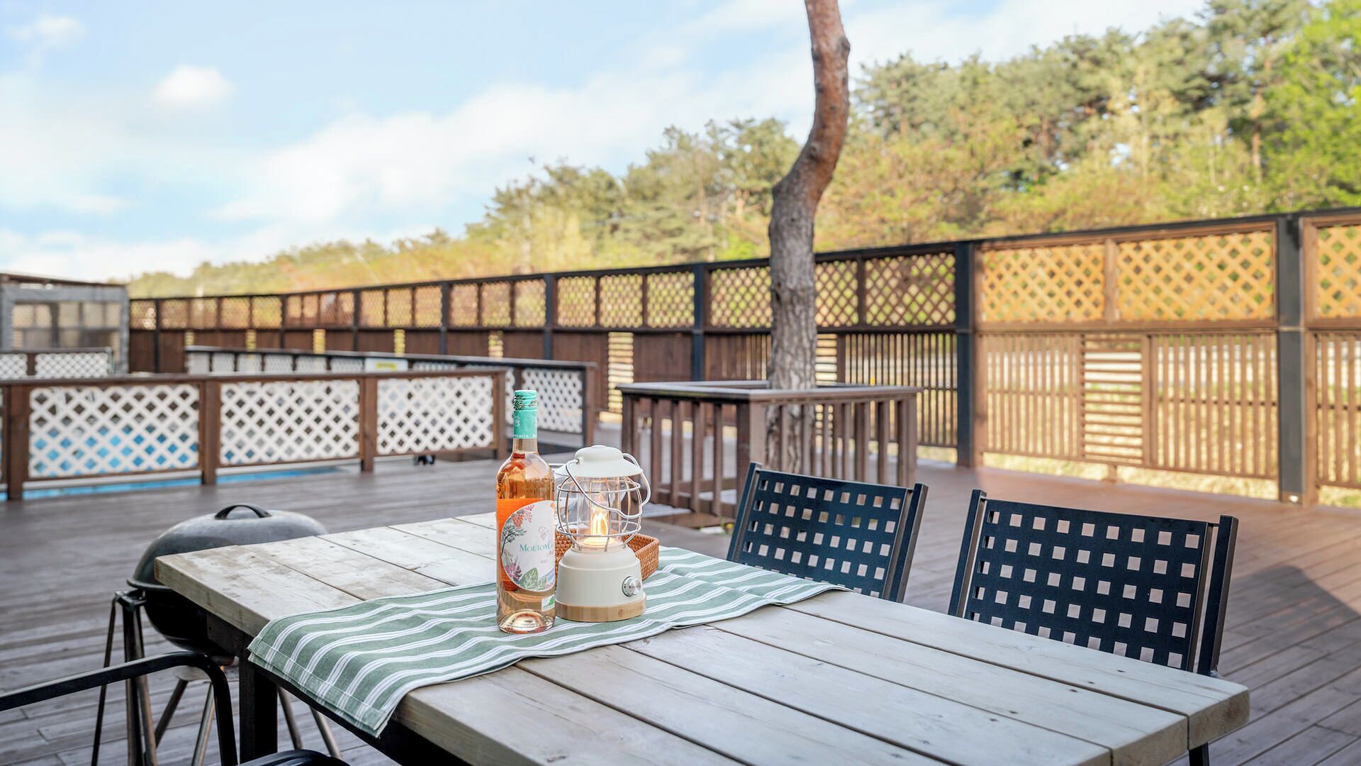 Une terrasse extérieure avec une table en bois, des chaises, une bouteille d’alcool, une lanterne et un barbecue.