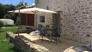 An outdoor patio with a stone wall, a table with chairs, and a large umbrella providing shade. - Vendée country cottage 5 minutes from La Roche sur Yon (La Genétouze)