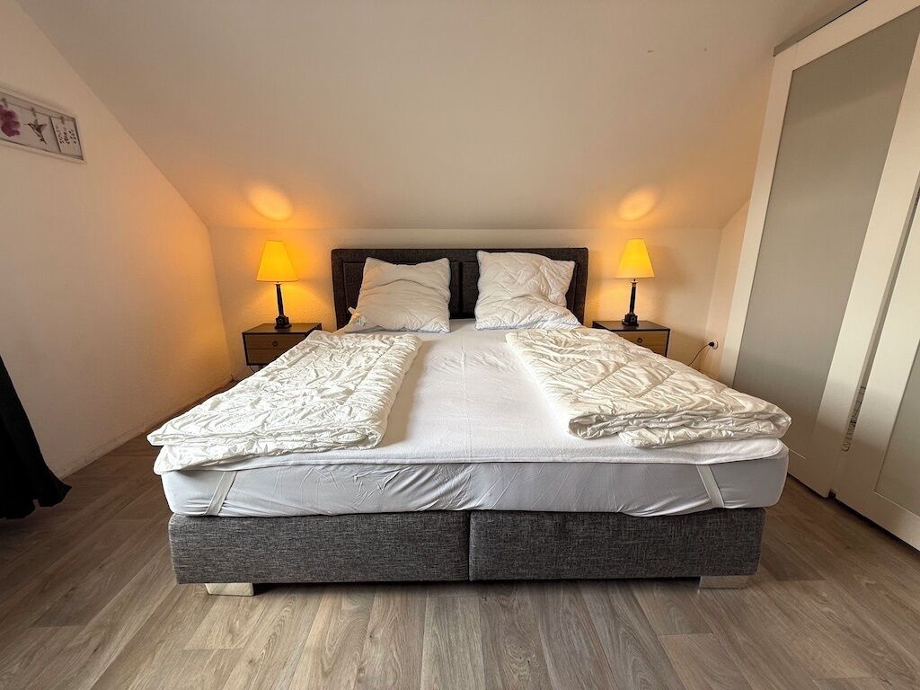 A neatly made double bed with a grey headboard and footboard, flanked by two bedside tables with lamps, in a room with wooden flooring and a door on the right.